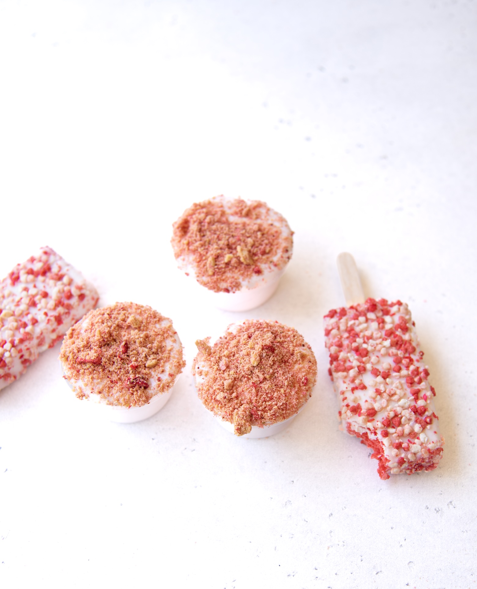 A group of five frozen strawberry-themed treats is arranged on a light, speckled surface. Three of the items are round pudding shots topped with a pinkish-brown crumb mixture, resembling crushed cookies and freeze-dried strawberries. Two rectangular strawberry shortcake ice cream bars, coated in a textured layer of white and red crumbs, are also included—one with a bite taken out. The overall presentation is clean, minimal, and bright, emphasizing the textures and colors of the desserts.