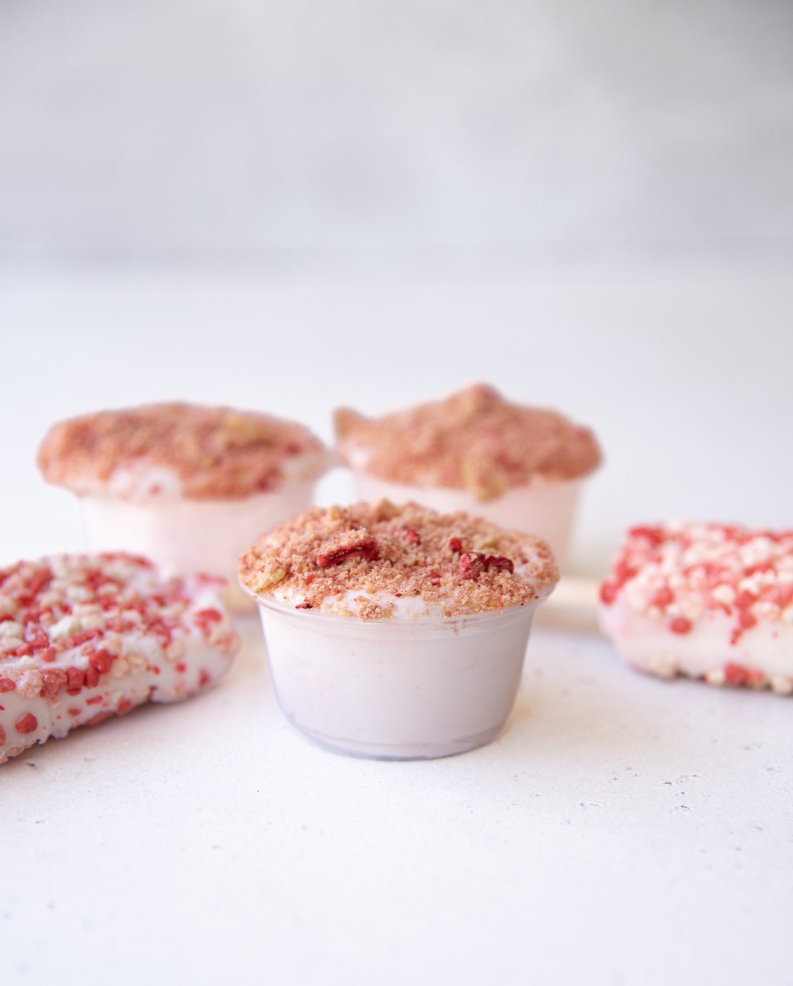 A close-up image shows five strawberry-themed desserts arranged on a light-colored surface. In the center is a clear plastic cup filled with pink pudding and topped with a crumbly pink and tan topping, resembling crushed cookies and freeze-dried strawberries. Surrounding it are four more similar pudding cups and two partially visible strawberry shortcake ice cream bars coated in red and white crumbs.