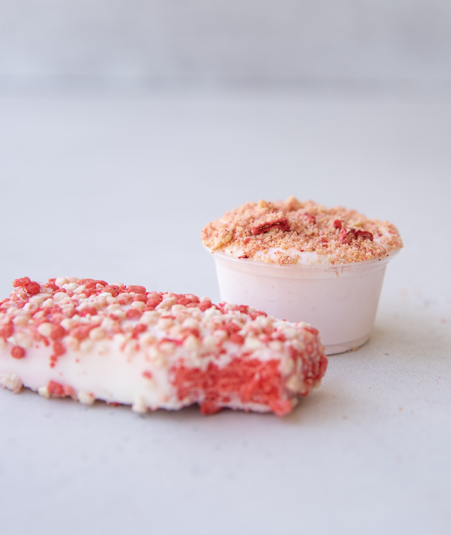 A close-up image features a single strawberry shortcake pudding shot and a strawberry shortcake ice cream bar. The pudding shot is in a small clear plastic cup filled with pale pink pudding and topped with a crumbly mixture of crushed cookies and freeze-dried strawberries. Next to it, the rectangular ice cream bar is coated in a textured layer of red and white crumbs, with a bite taken out, revealing the vibrant red strawberry center.