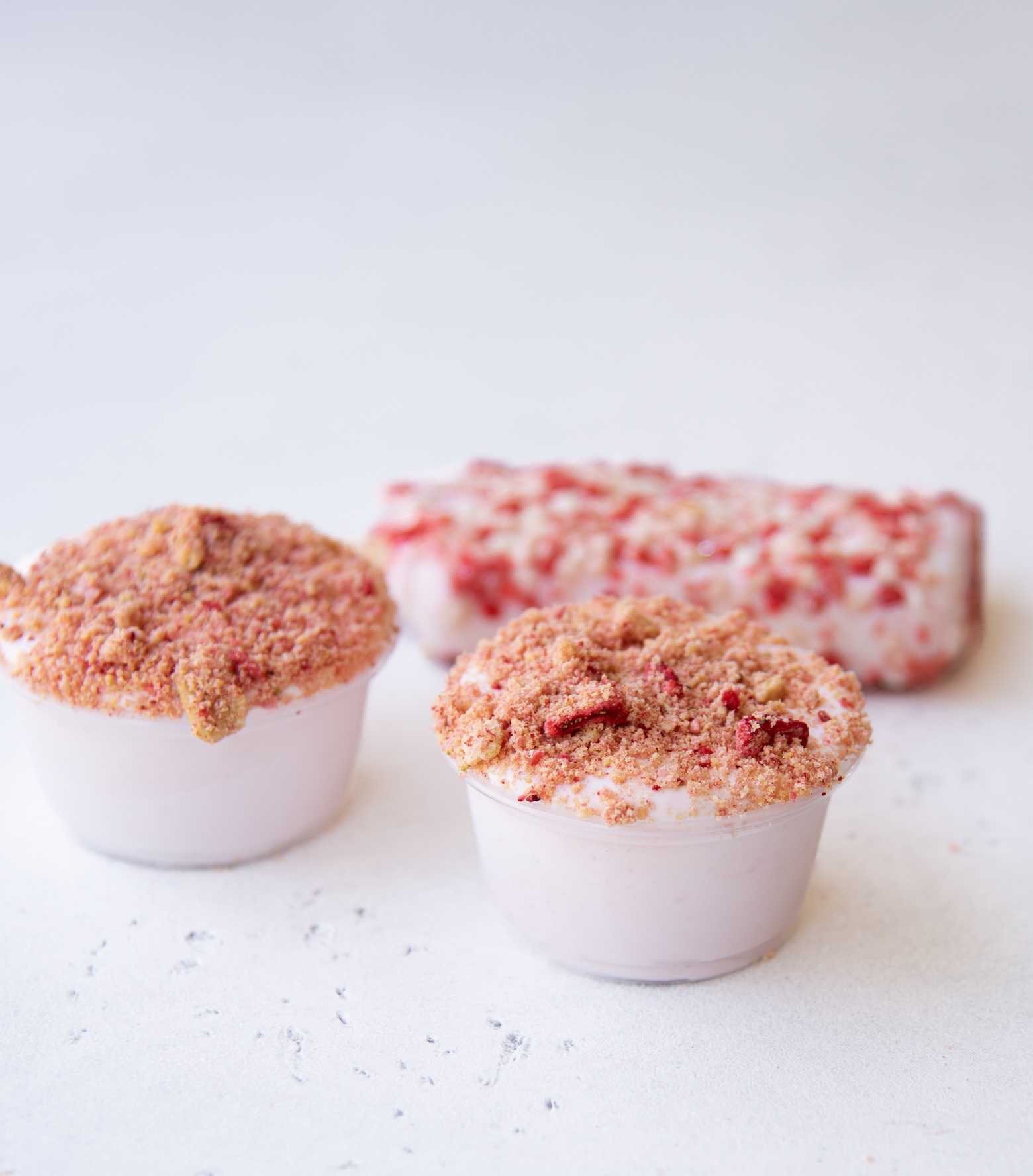Two strawberry shortcake pudding shots sit in the foreground of a light-colored surface. Each shot is in a small clear plastic cup filled with pale pink pudding and topped with a generous layer of crumbled cookie and freeze-dried strawberry topping. In the background, a single strawberry shortcake ice cream bar lies slightly out of focus, coated in a textured mix of red and white crumbs.