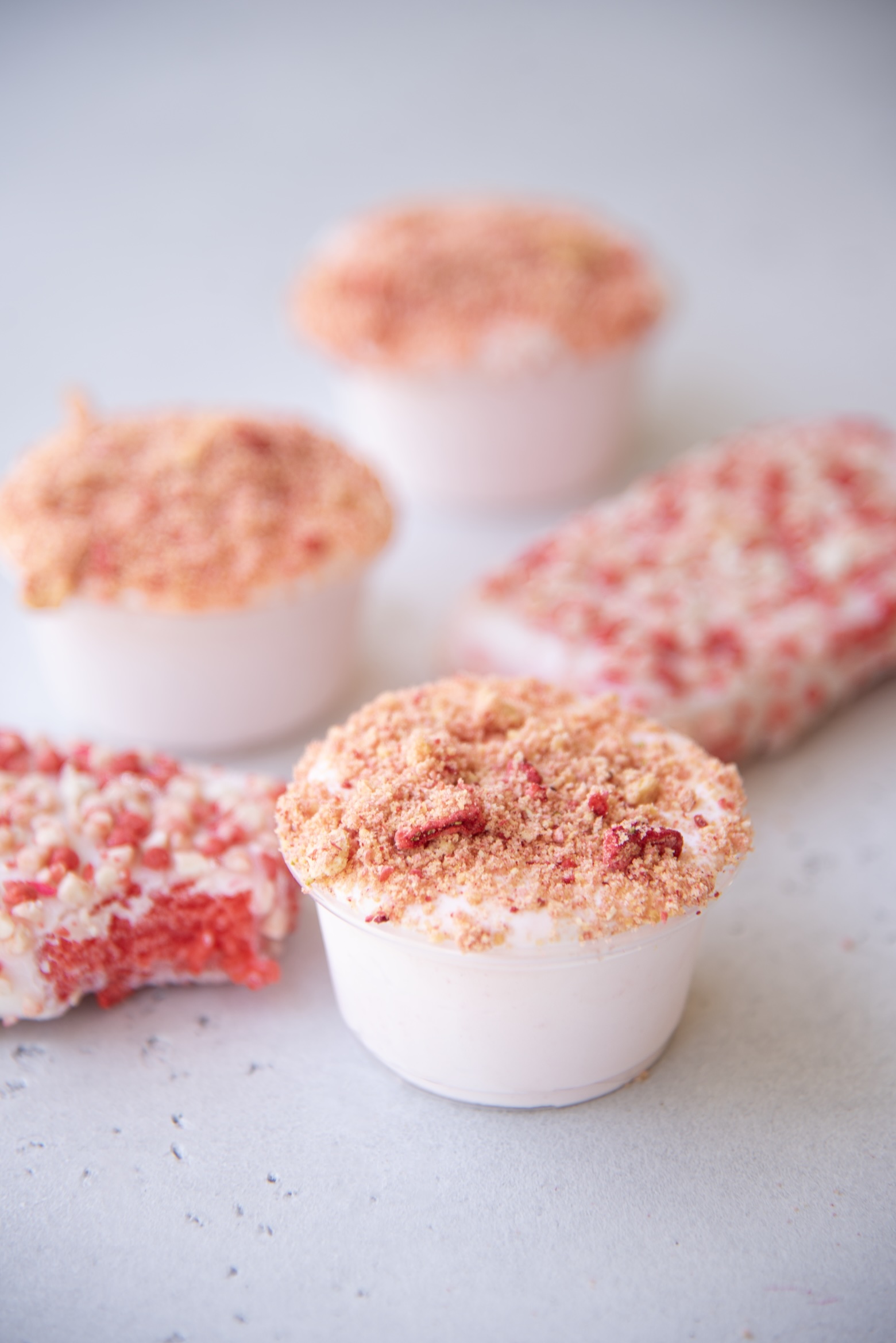 A close-up image showcases a group of strawberry shortcake pudding shots and ice cream bars on a light gray surface. In the foreground, a single pudding shot in a small clear cup is topped with a generous layer of pink and tan crumb topping made of crushed cookies and freeze-dried strawberries. Surrounding it are more pudding shots and two rectangular strawberry shortcake ice cream bars coated in red and white crumbs, one with a bite taken out.