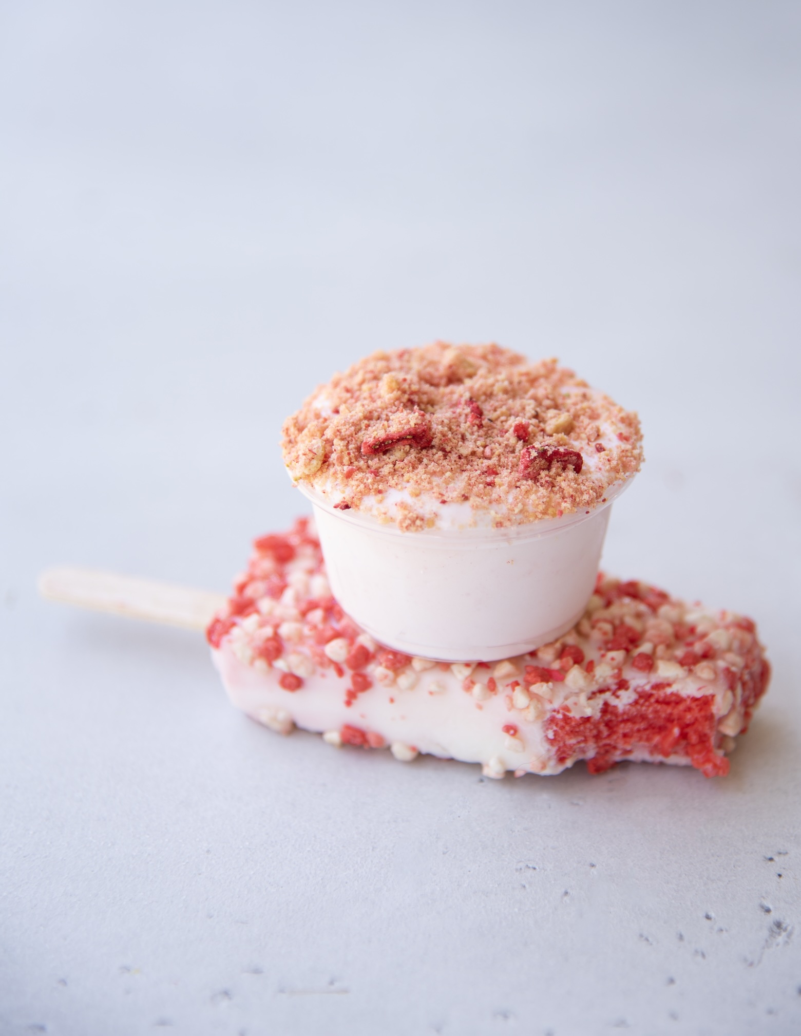 A strawberry shortcake pudding shot is stacked on top of a partially eaten strawberry shortcake ice cream bar. The pudding shot is in a small clear plastic cup, filled with light pink pudding and topped with a crumbly mixture of crushed cookies and freeze-dried strawberries. The ice cream bar underneath has a textured coating of red and white crumbs and a visible bite revealing a bright red interior. The treats rest on a smooth, pale gray surface with a soft gradient background.