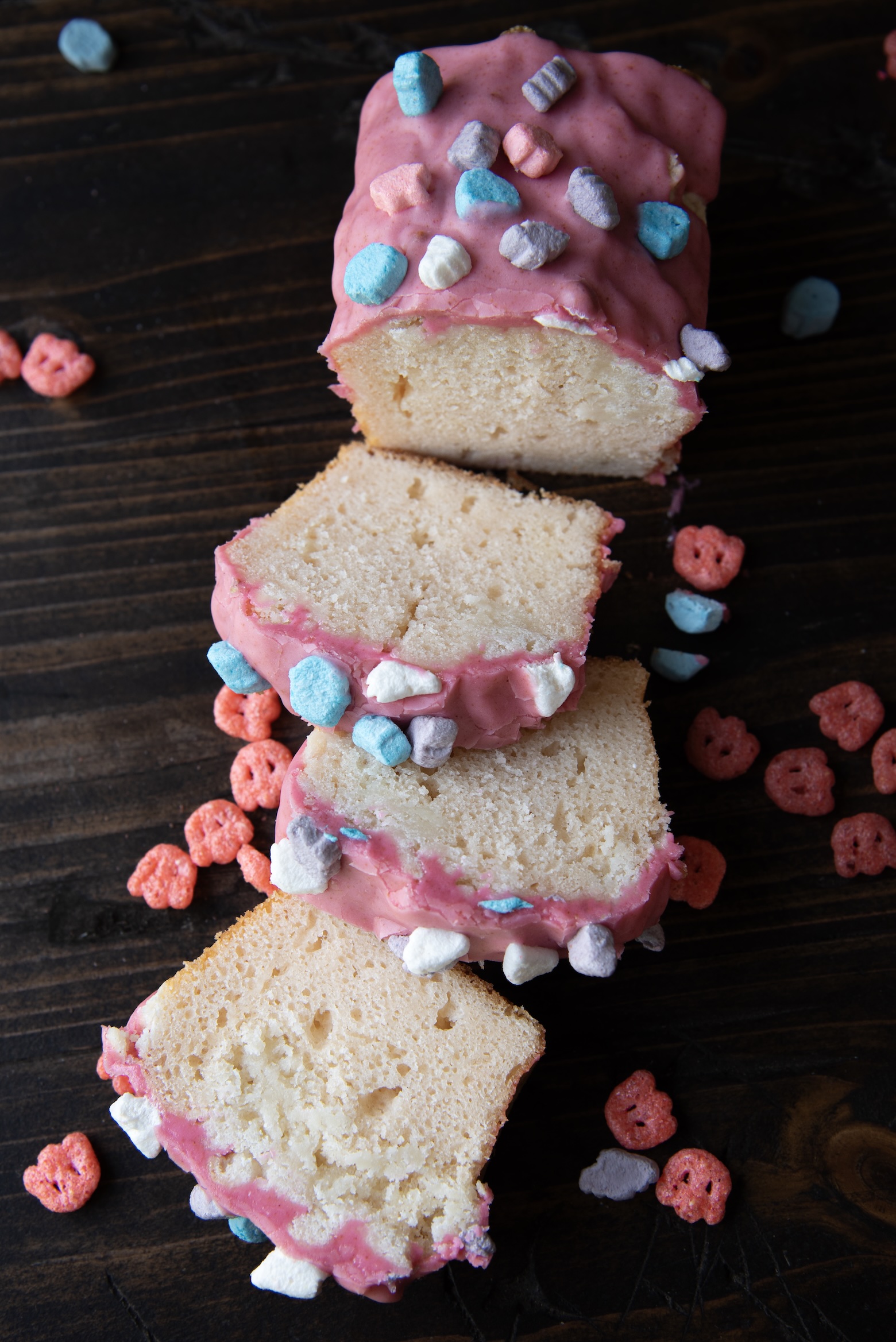 A loaf of Franken Berry pound cake is sliced and arranged on a dark wooden surface. The cake is topped with a thick layer of glossy pink frosting and sprinkled with colorful cereal marshmallows in shades of blue, purple, pink, and white. A few marshmallows and red cereal pieces are scattered around the cake slices, adding a playful, nostalgic touch. The inside of the cake appears dense and moist, contrasting with the bright, whimsical topping.