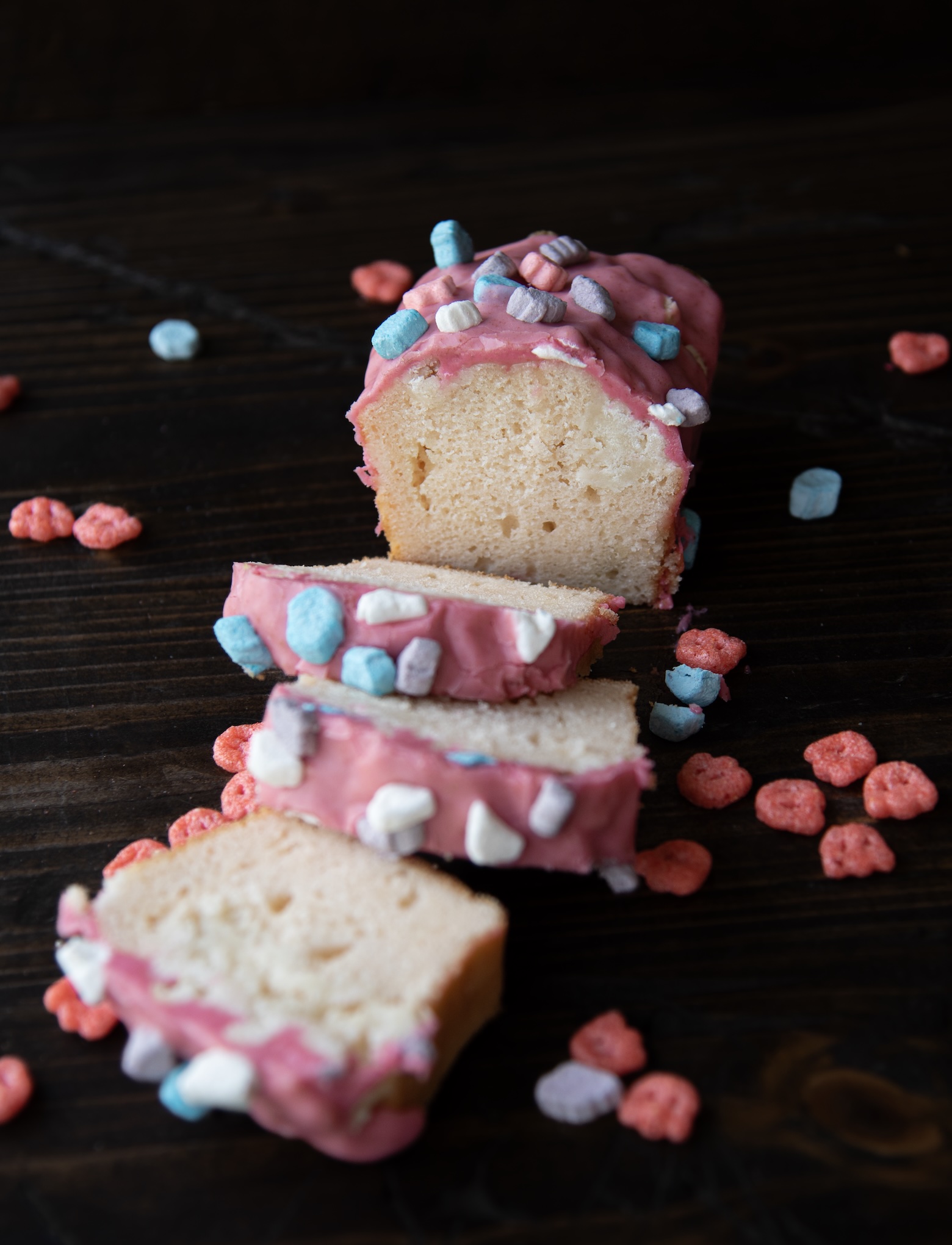 A loaf of Franken Berry pound cake with pink frosting is sliced and displayed on a dark wooden surface. The frosting is thick and creamy, dripping slightly over the edges and topped with colorful cereal marshmallows in shades of blue, purple, pink, and white. Red cereal pieces are scattered playfully around the slices, creating a fun, nostalgic scene reminiscent of childhood breakfast treats. The inside of the cake looks moist and tender, contrasting with the bright, candy-like topping.