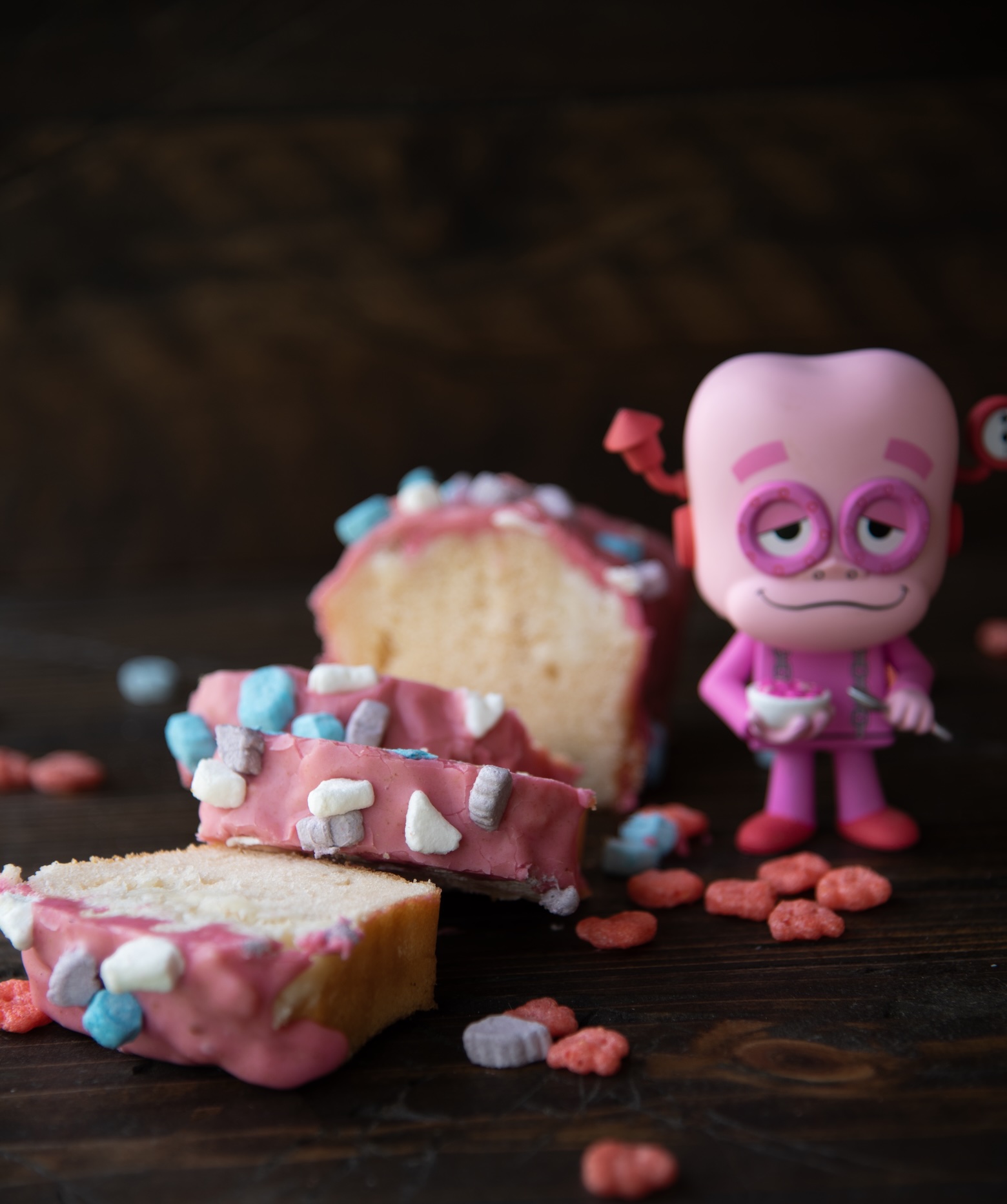 A sliced loaf of Franken Berry pound cake with pink frosting and colorful cereal marshmallows is arranged on a dark wooden surface. Scattered red cereal pieces surround the slices. To the right of the cake stands a small pink figurine of Franken Berry, the classic cereal mascot, with a sleepy expression and one raised eyebrow, holding a tiny white bowl. The scene has a playful, nostalgic tone, combining the dessert and the cereal character.