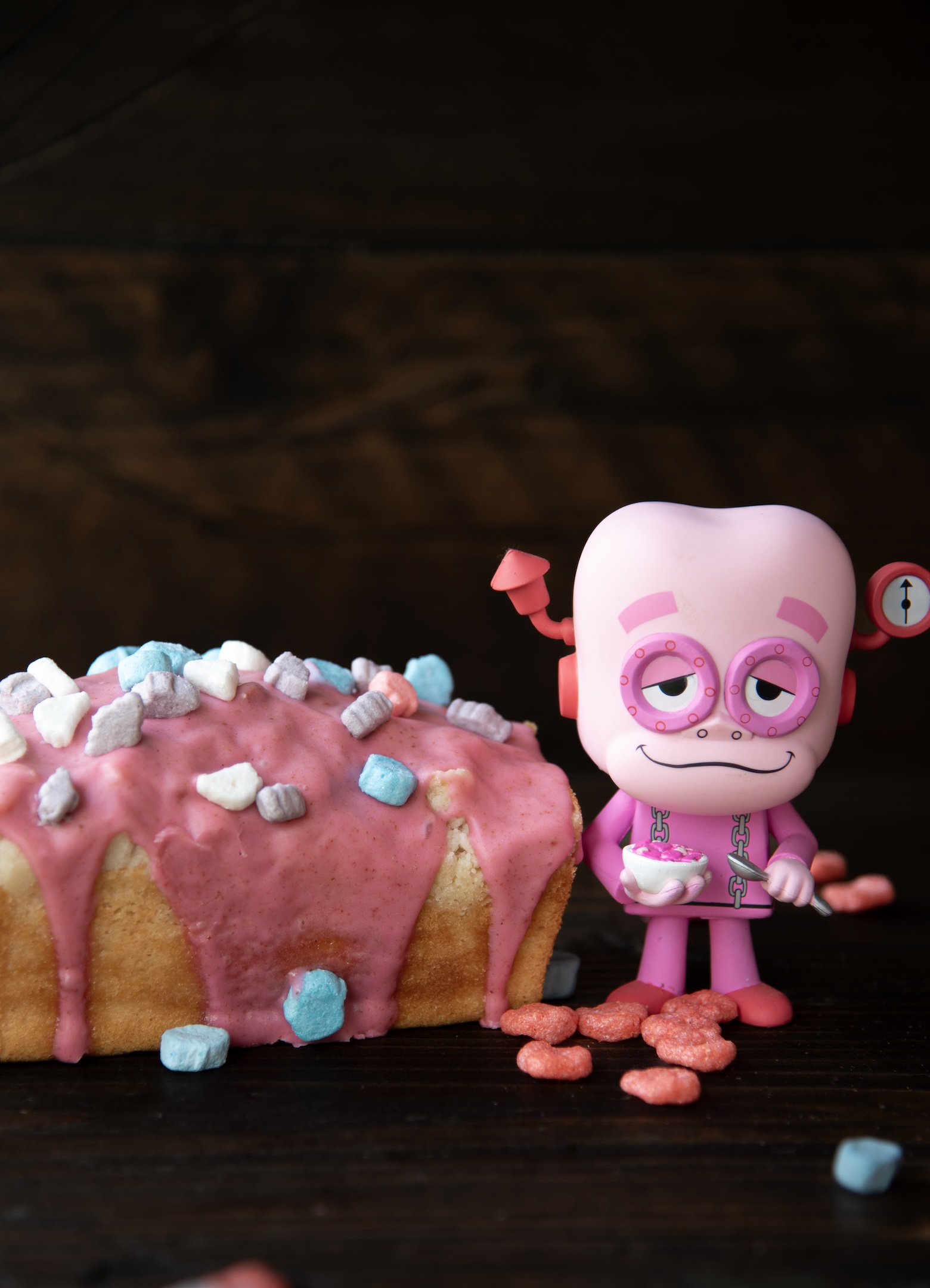 A small loaf cake with thick pink frosting and colorful cereal marshmallows sits on a dark wooden surface. Beside it stands a pink Franken Berry figurine holding a tiny bowl and spoon, smiling with a sleepy, mischievous expression. Red cereal pieces are scattered around the figure and cake, adding a playful, nostalgic breakfast cereal theme to the scene. The frosting drips slightly down the sides of the golden cake, creating a fun, whimsical presentation.