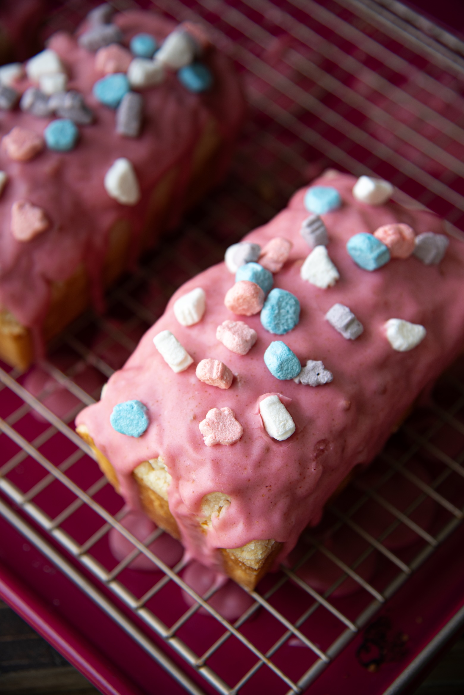 Two small loaf cakes rest on a wire cooling rack set over a magenta tray. Each loaf is coated in a thick, glossy pink glaze that drips slightly down the sides. The tops are decorated with colorful cereal marshmallows in pastel shades of blue, pink, purple, and white, creating a fun and whimsical look reminiscent of breakfast cereal treats. The bright frosting and marshmallows contrast with the golden color of the cake underneath.