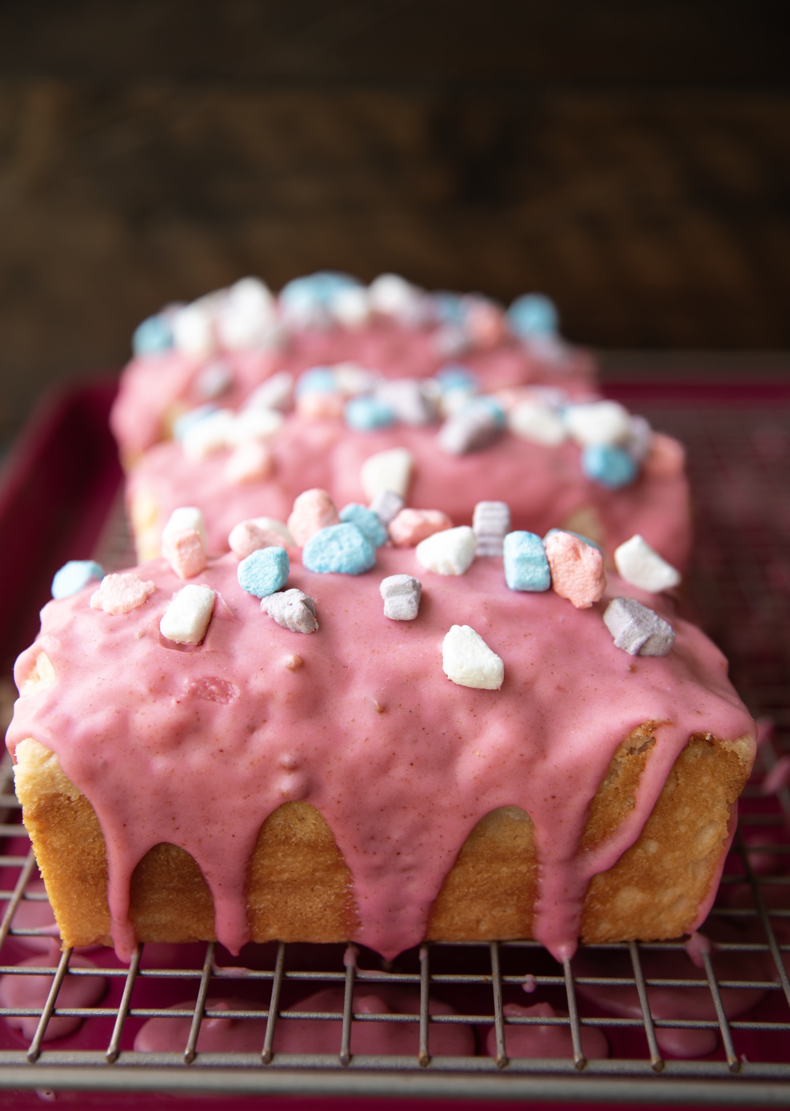 Three small loaf cakes are lined up on a wire cooling rack over a magenta tray. Each cake is coated with a thick, glossy layer of pink frosting that drips slightly down the golden edges. The tops are sprinkled with colorful cereal marshmallows in shades of blue, white, pink, and purple, adding a whimsical, nostalgic touch. The frosting’s smooth sheen and the pastel colors of the marshmallows create a playful, dessert-inspired scene reminiscent of classic breakfast cereals.