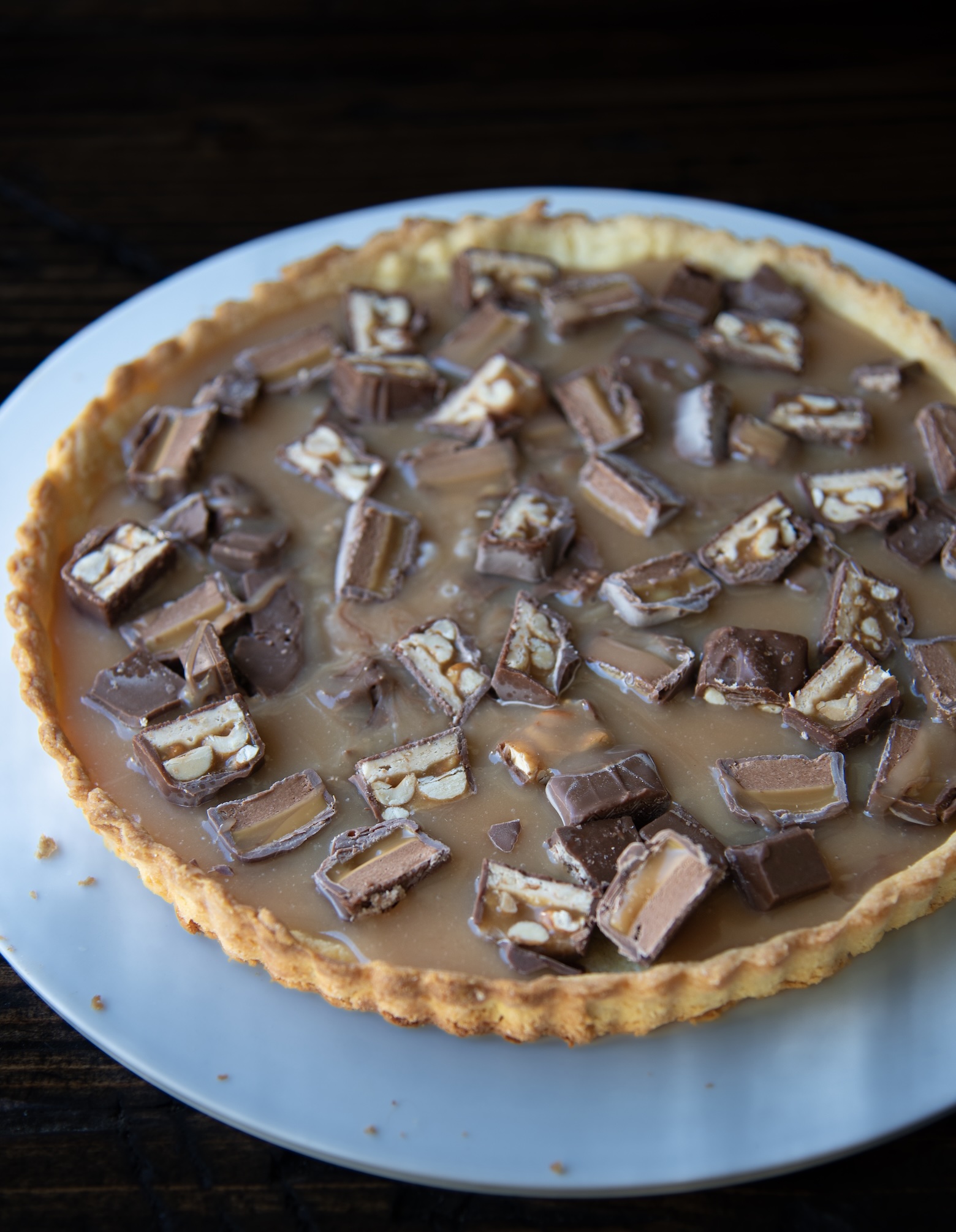 Alt text: A caramel tart with a golden, flaky crust topped with chopped Snickers candy bars. The glossy caramel filling is studded with chunks of chocolate showing layers of nougat, peanuts, and caramel. The tart sits on a white plate atop a dark wooden surface, with a few crumbs scattered near the edge.