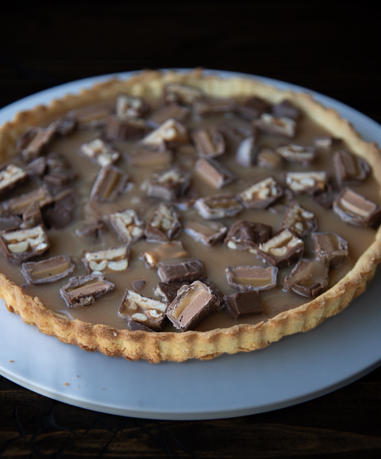 Alt text: A close-up view of a caramel tart topped with chopped pieces of Snickers candy bars. The tart has a golden-brown, flaky crust filled with glossy caramel and generously scattered chunks of chocolate showing layers of nougat, caramel, and peanuts. The tart rests on a white plate, with a dark wooden surface beneath, emphasizing the rich textures and warm tones of the dessert.