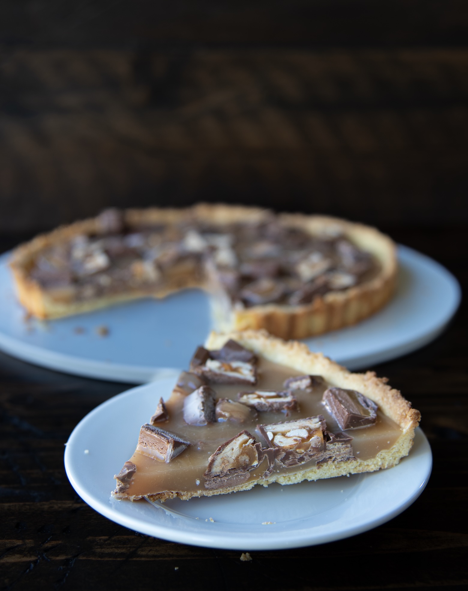 A slice of caramel tart topped with chopped Snickers candy pieces sits on a small white plate in the foreground. Behind it, the remaining tart with a missing slice rests on a larger white plate. The tart has a golden, crumbly crust and a glossy caramel filling studded with chocolate pieces showing layers of nougat, peanuts, and caramel. Both plates are placed on a dark wooden surface with a softly blurred background.