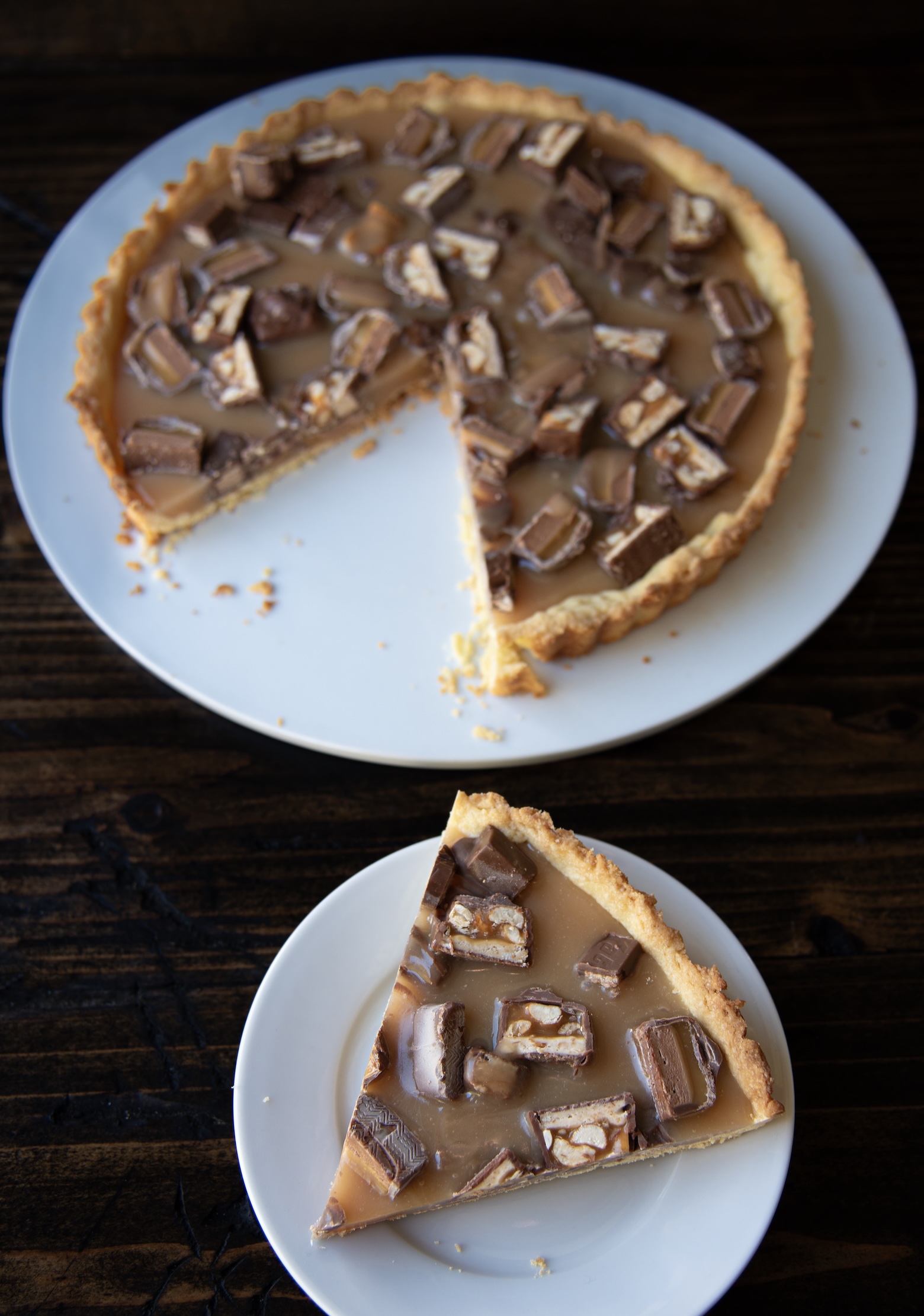 Alt text: A caramel tart topped with chopped Snickers candy pieces sits on a white plate. One slice has been removed and placed on a smaller white plate in the foreground. The tart has a golden, crumbly crust filled with smooth caramel and generous chunks of chocolate bars showing layers of nougat, peanuts, and caramel. Both plates rest on a dark wooden surface, highlighting the glossy caramel and textures of the candy pieces.