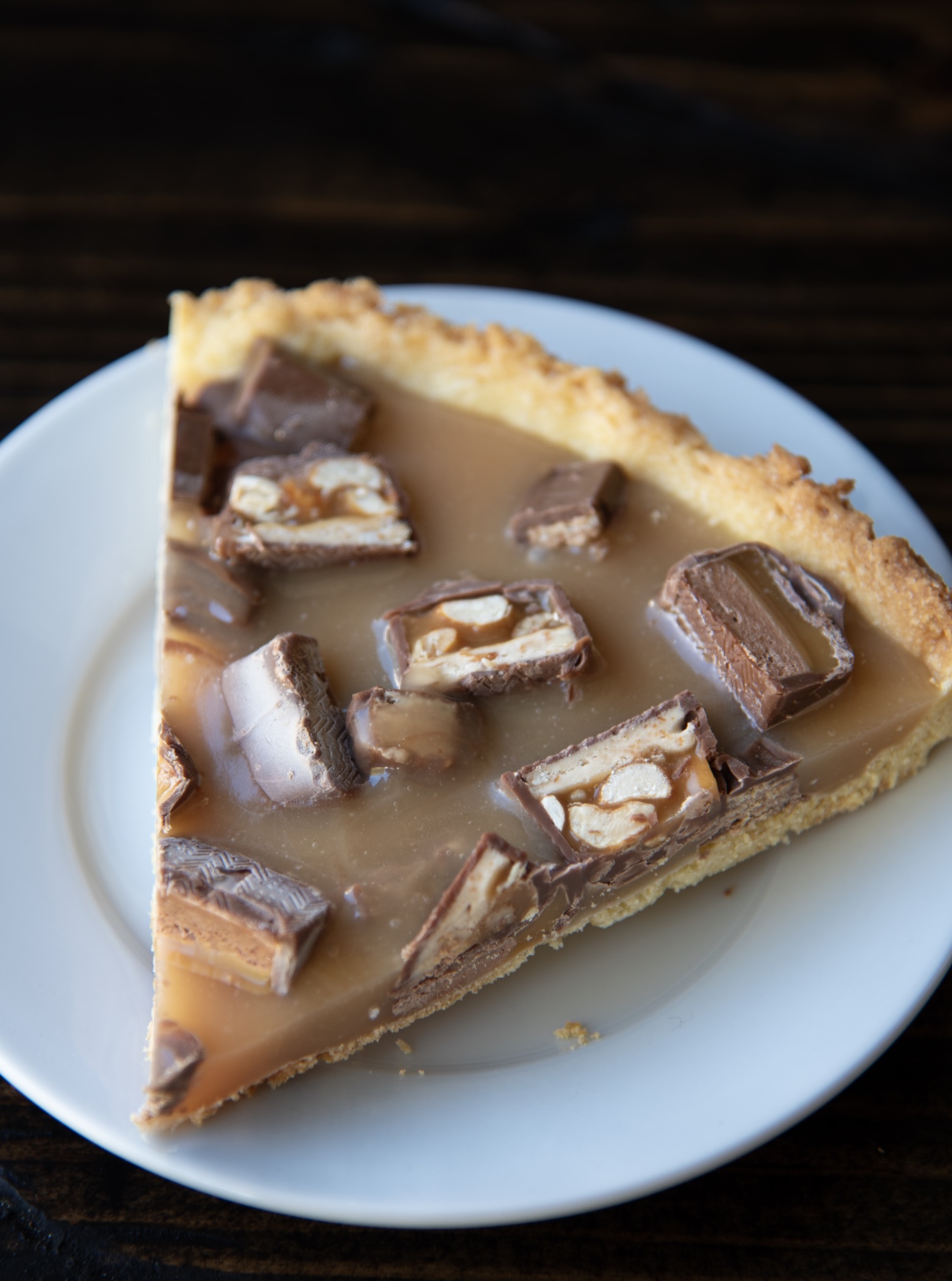 A close-up overhead view of a single slice of caramel tart on a white plate. The tart slice has a golden, crumbly crust and is filled with smooth caramel topped with chopped Snickers pieces showing layers of chocolate, nougat, peanuts, and caramel. The glossy caramel surface contrasts with the textured candy chunks, and the plate rests on a dark wooden table.