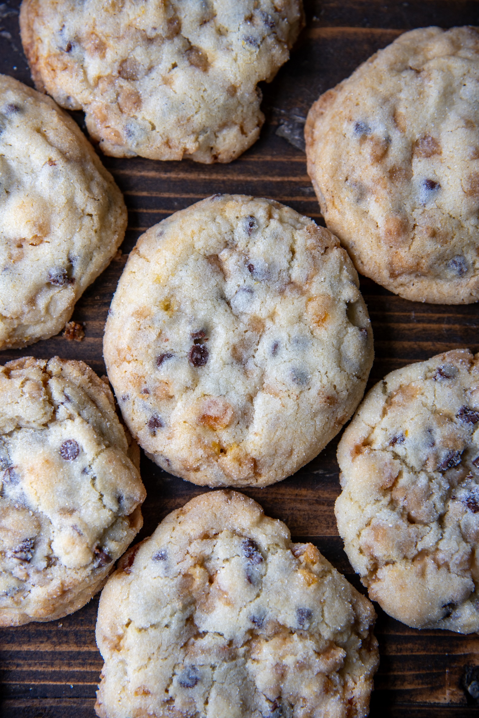 Close up of freshly baked chocolate chip cookies with golden edges and soft centers, dotted with chocolate chips and small tangerine bits, arranged on a rustic wooden surface.