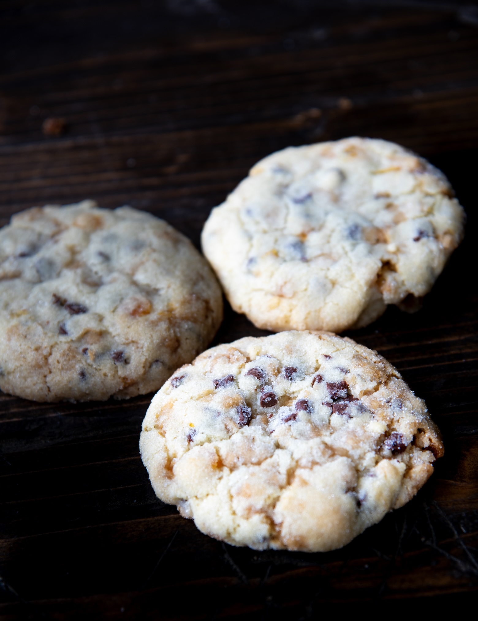 Close up of two soft, thick chocolate chip cookies with a slightly crumbly texture and golden edges, resting on a dark wooden surface with the background softly blurred.