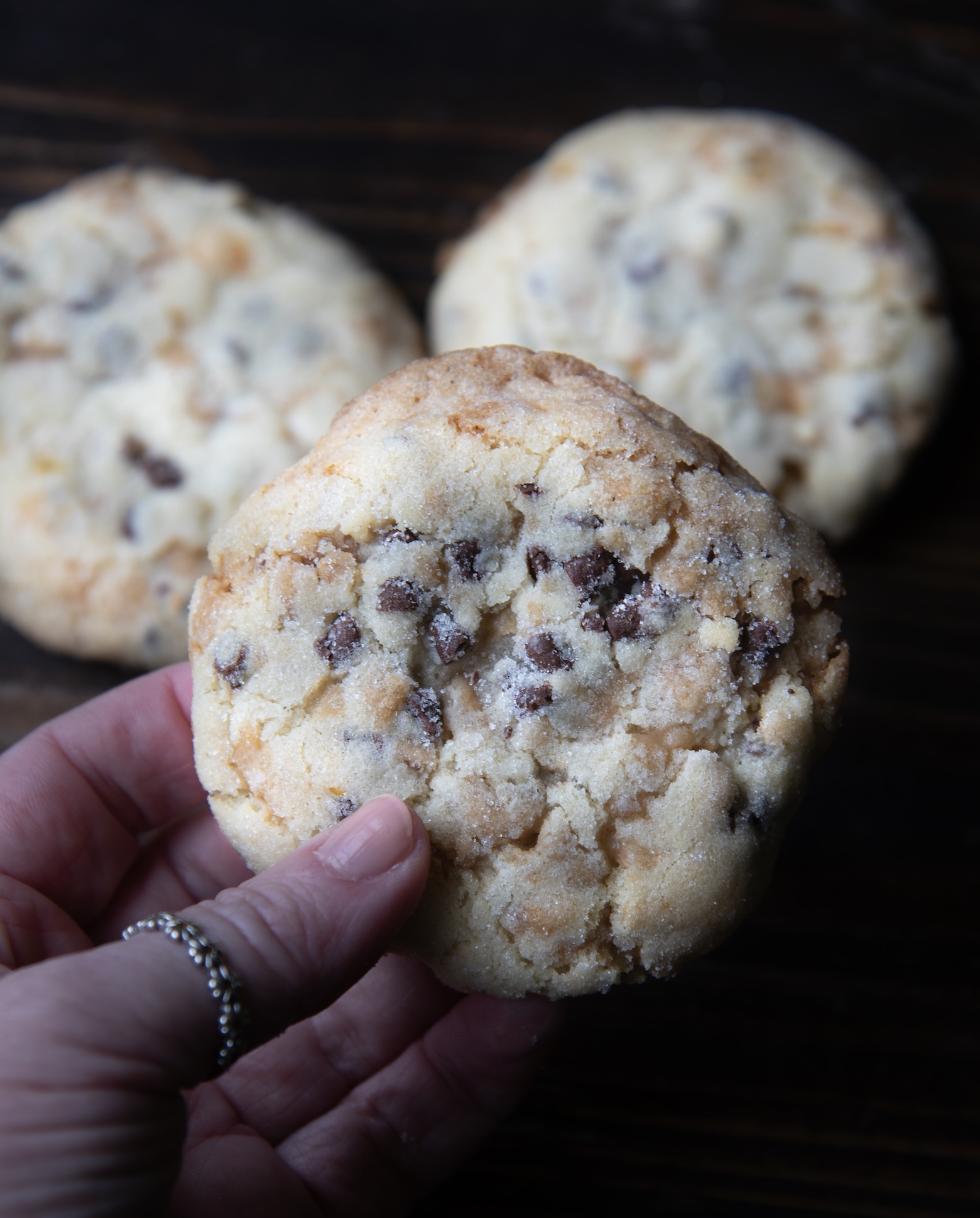 Hand holding a thick, soft chocolate chip cookie with a crumbly texture and golden edges, with two more cookies blurred in the background on a dark wooden surface.