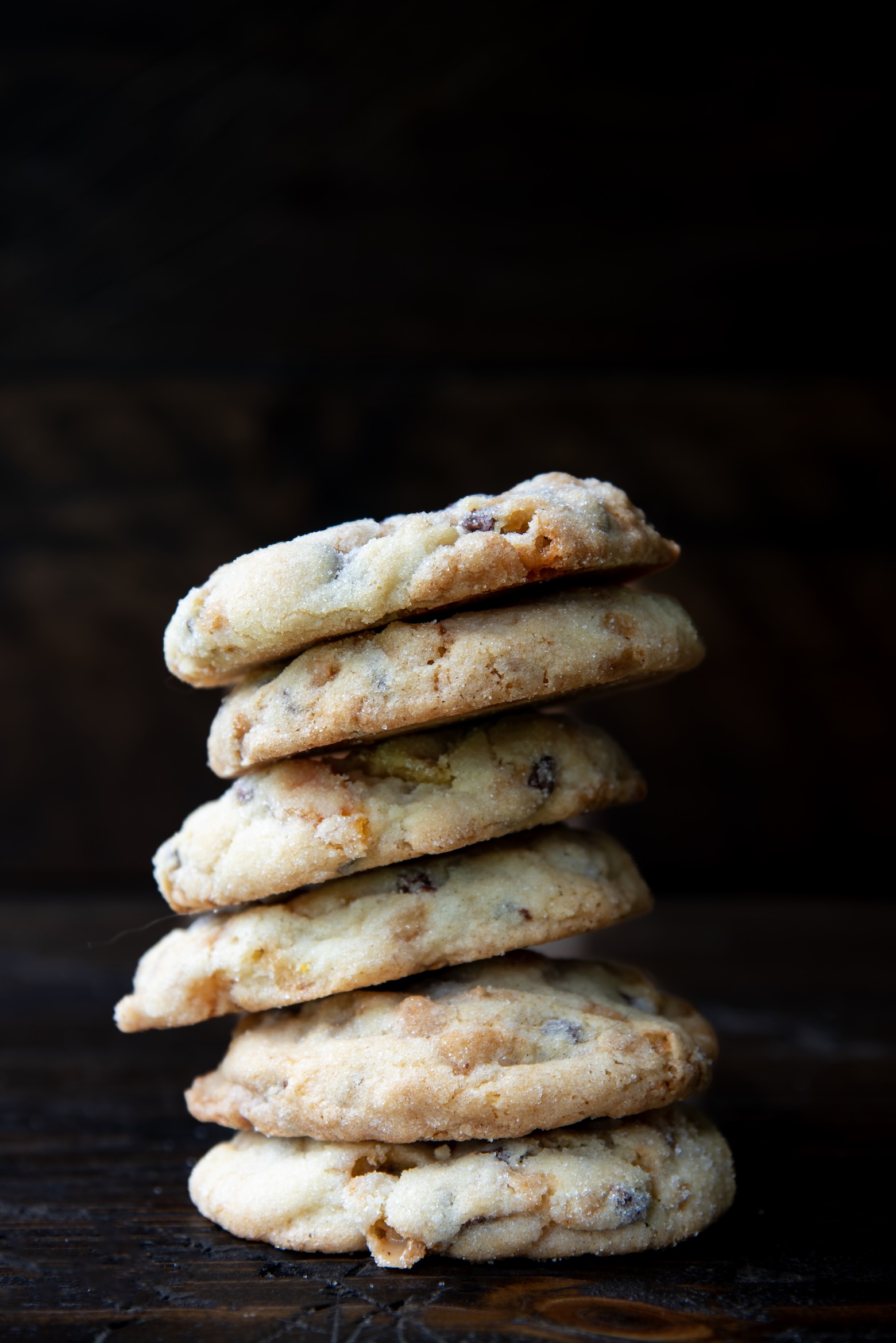 Stack of thin, golden chocolate chip cookies with slightly crisp edges and a soft center, piled on a dark wooden surface against a dark background.