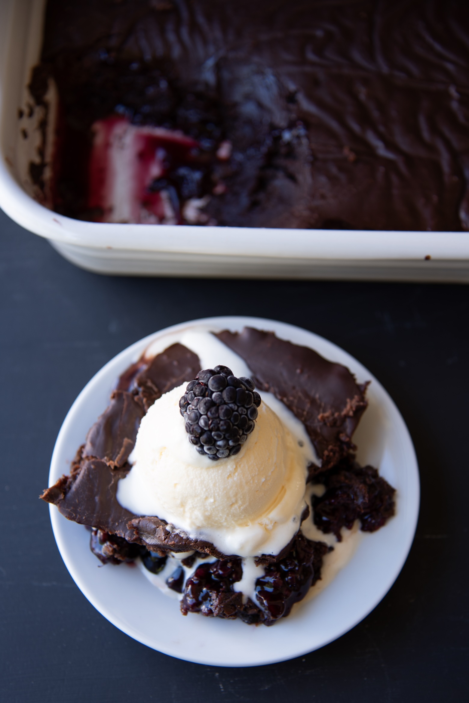 Serving of chocolate blackberry dump cake topped with vanilla ice cream and a fresh blackberry, with the baking dish in the background showing the rich, gooey berry filling.
