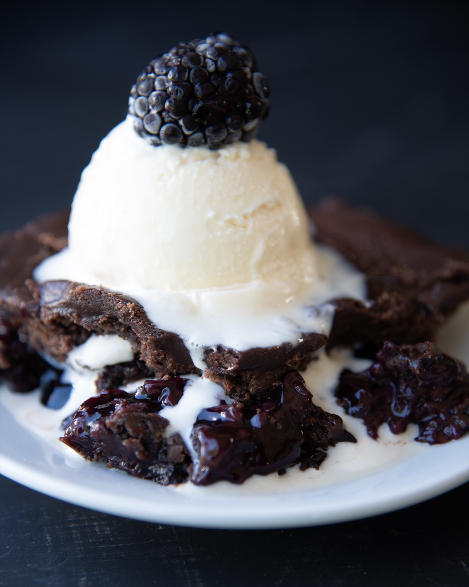 Close up of rich chocolate blackberry dump cake topped with a melting scoop of vanilla ice cream and a fresh blackberry, with glossy berry filling and chocolate edges peeking through.