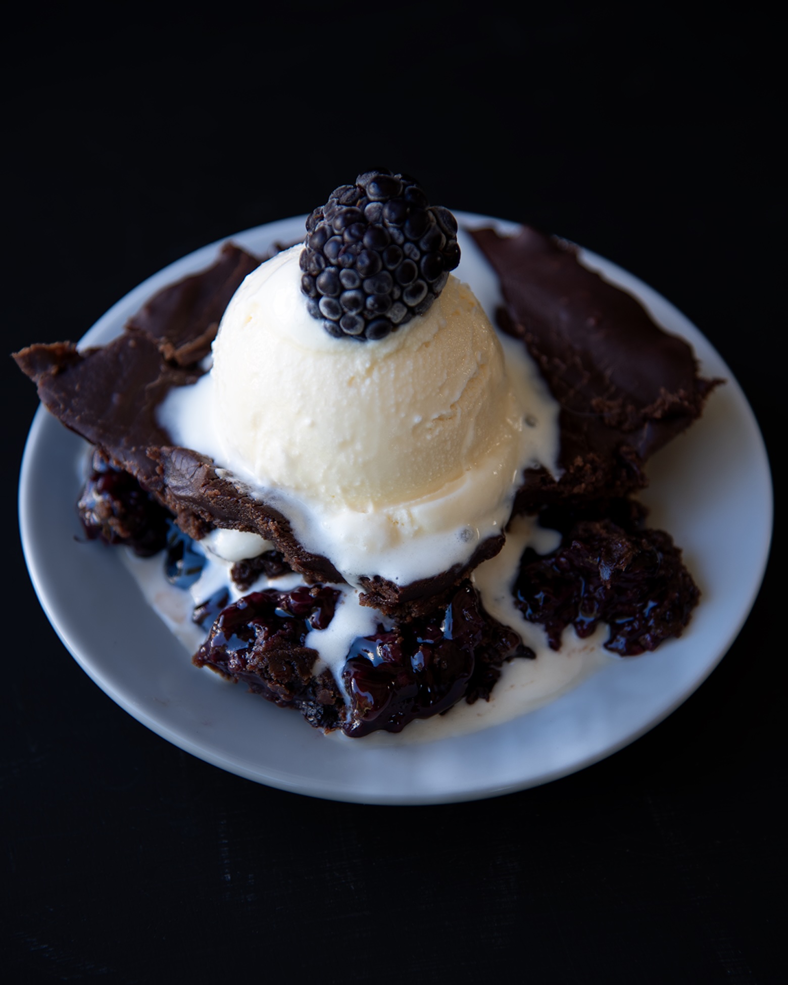 Chocolate blackberry dump cake on a white plate, topped with a scoop of melting vanilla ice cream and a fresh blackberry, with rich berry filling and chocolate edges visible against a dark background.