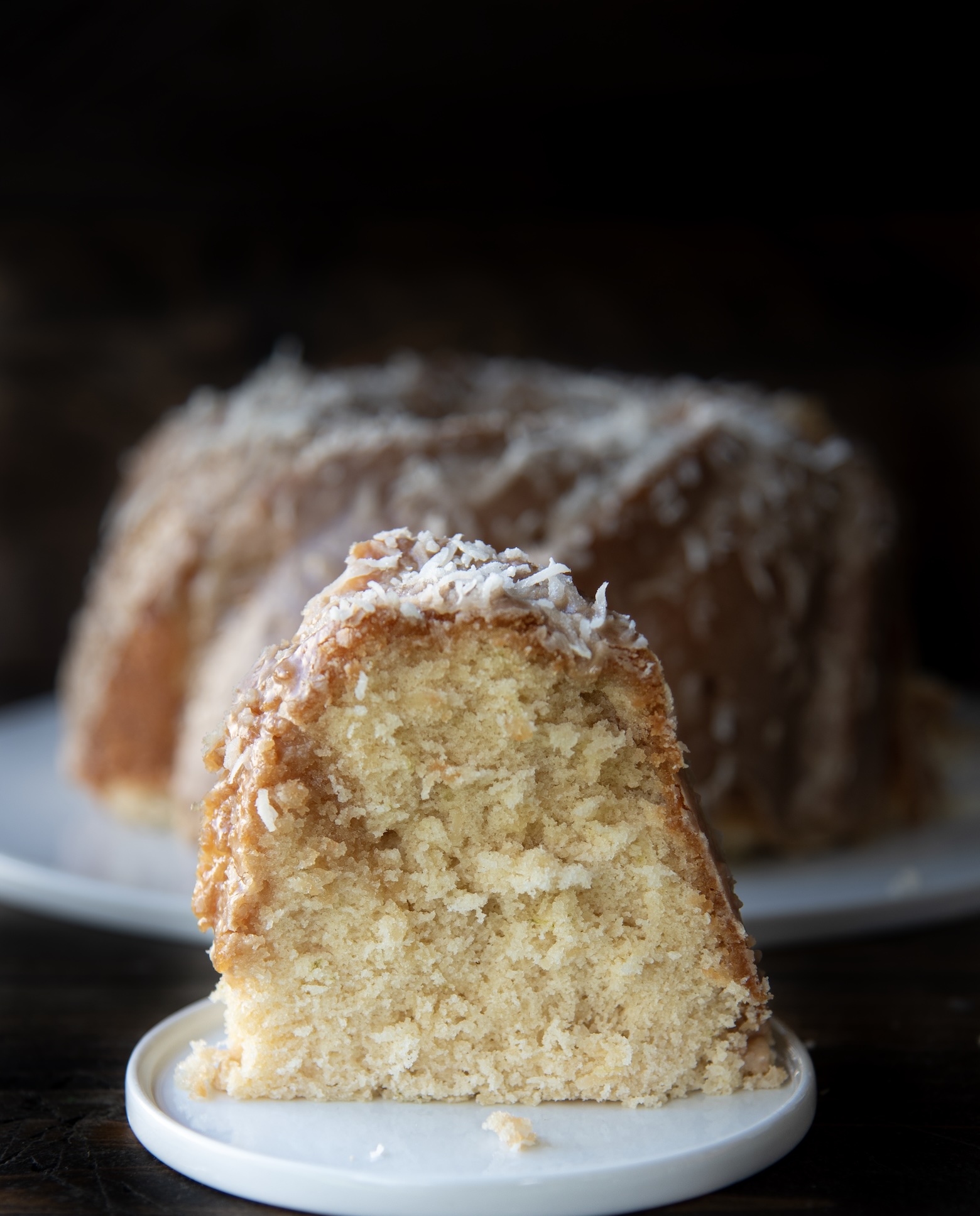 Slice of pineapple coconut Bundt cake with coconut frosting and shredded coconut, served in front of the whole cake.