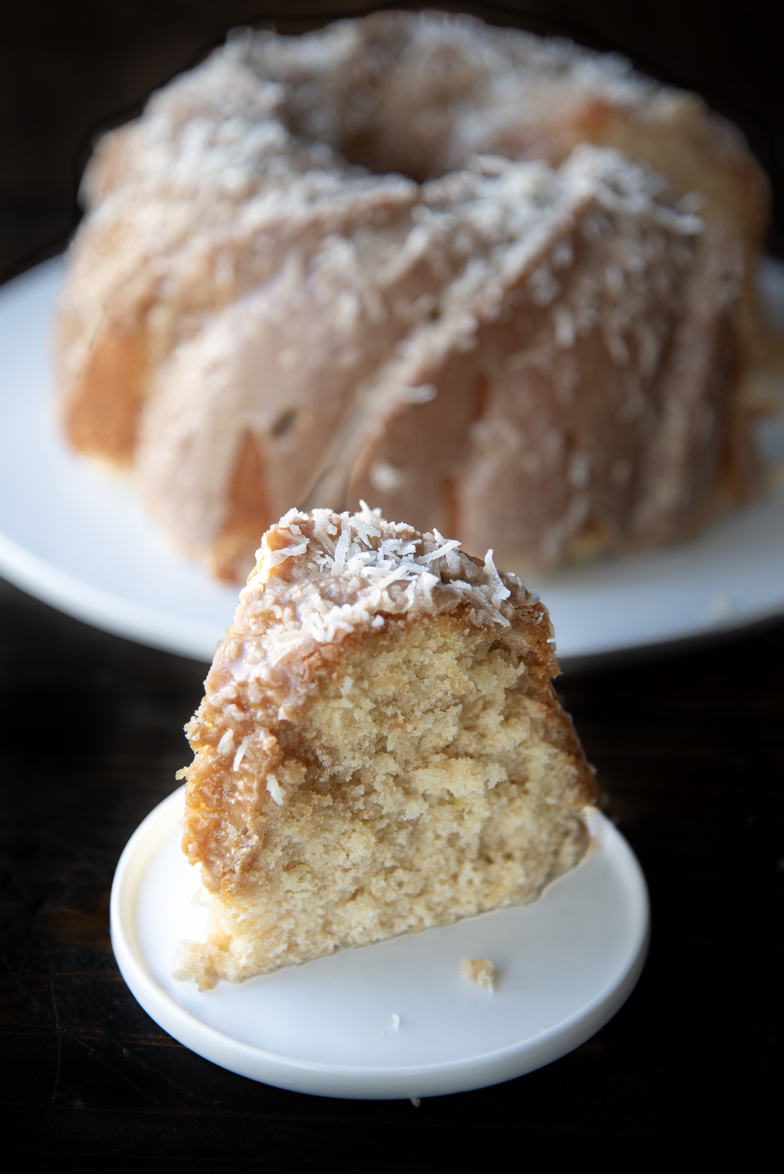 Slice of pineapple coconut Bundt cake topped with coconut frosting and shredded coconut, with the whole cake blurred in the background.